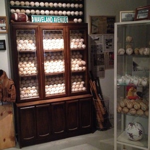 May include: A dark wood cabinet with glass doors displays a collection of baseballs. The cabinet has four drawers below the glass doors. A sign above the cabinet reads "Waveland Avenue".