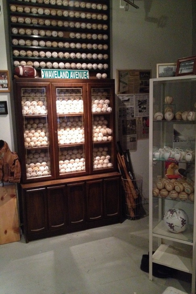 May include: A dark wood cabinet with glass doors displays a collection of baseballs. The cabinet has four drawers below the glass doors. A sign above the cabinet reads "Waveland Avenue".