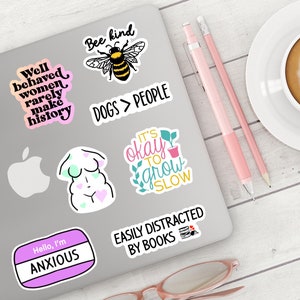 May include: A laptop computer with several colourful stickers on it. The stickers feature various phrases and images, including "Well behaved women rarely make history", "Bee kind", "Dogs > People", "It's okay to grow slow", "Easily distracted by books", and "Hello, I'm Anxious".