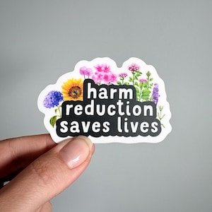 May include: A die-cut sticker featuring a black rectangular shape with rounded corners and the white text "harm reduction saves lives" printed in a bold sans-serif font, surrounded by colorful illustrations of various wildflowers.