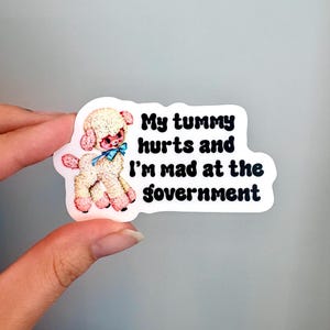 May include: A die-cut sticker featuring a vintage-style illustration of a cream-colored lamb with pink accents and a blue bow. The text printed in black, bold, rounded lettering reads: My tummy hurts and I'm mad at the government.