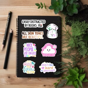 May include: A collection of colorful stickers with various phrases and illustrations on a black notebook. The stickers include text like "Easily Distracted by Books," "All Skin Tones Are Beautiful," and "Nice People Use Drugs." Other stickers feature a cloud, a body outline, and plants.
