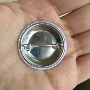May include: The back of a silver button with a white border. A safety pin is attached to the back of the button.