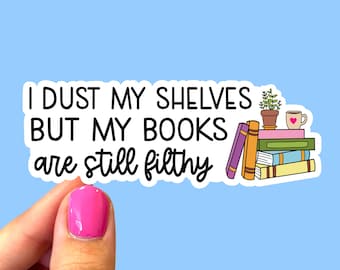 May include: A white sticker with black text that reads "I dust my shelves but my books are still filthy". The sticker features a cartoon illustration of a stack of books with a potted plant and a cup of coffee on top.