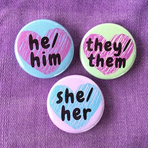 May include: Three round buttons with pink and blue hearts and the words "he/him", "she/her", and "they/them" in black.