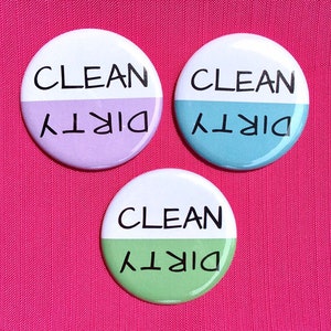Könnte beinhalten: Drei runde Knöpfe mit weißem Rücken, auf denen die Wörter "CLEAN" und "DIRTY" in Schwarz auf gegenüberliegenden Seiten gedruckt sind. Die Knöpfe sind in der Mitte geteilt, wobei jede Hälfte eine andere Farbe hat: lila, blau und grün.