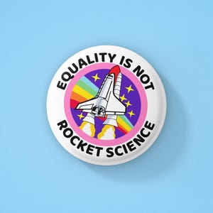 Peut inclure: Bouton blanc avec un cercle rose et le texte "EQUALITY IS NOT ROCKET SCIENCE" en noir. À l'intérieur du cercle se trouvent un arc-en-ciel, des étoiles et une navette spatiale blanche avec des accents rouges.