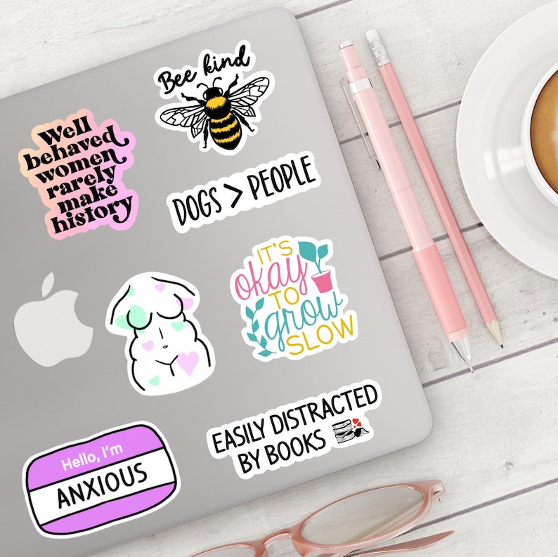May include: A laptop computer with several colorful stickers on it. The stickers feature various phrases and images, including "Well behaved women rarely make history", "Bee kind", "Dogs > People", "It's okay to grow slow", "Easily distracted by books", and "Hello, I'm Anxious".