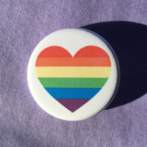 May include: A white button featuring a rainbow heart design. The heart is composed of six stripes: red, orange, yellow, green, blue, and purple.