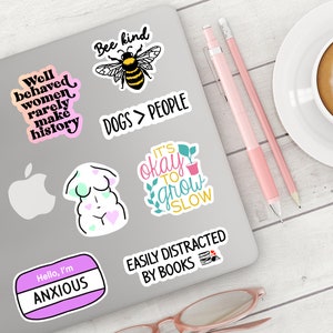 May include: A laptop computer with various stickers on it. The stickers feature phrases like "Well behaved women rarely make history", "Bee kind", "Dogs > People", "It's okay to grow slow", "Easily distracted by books", and "Hello, I'm Anxious".
