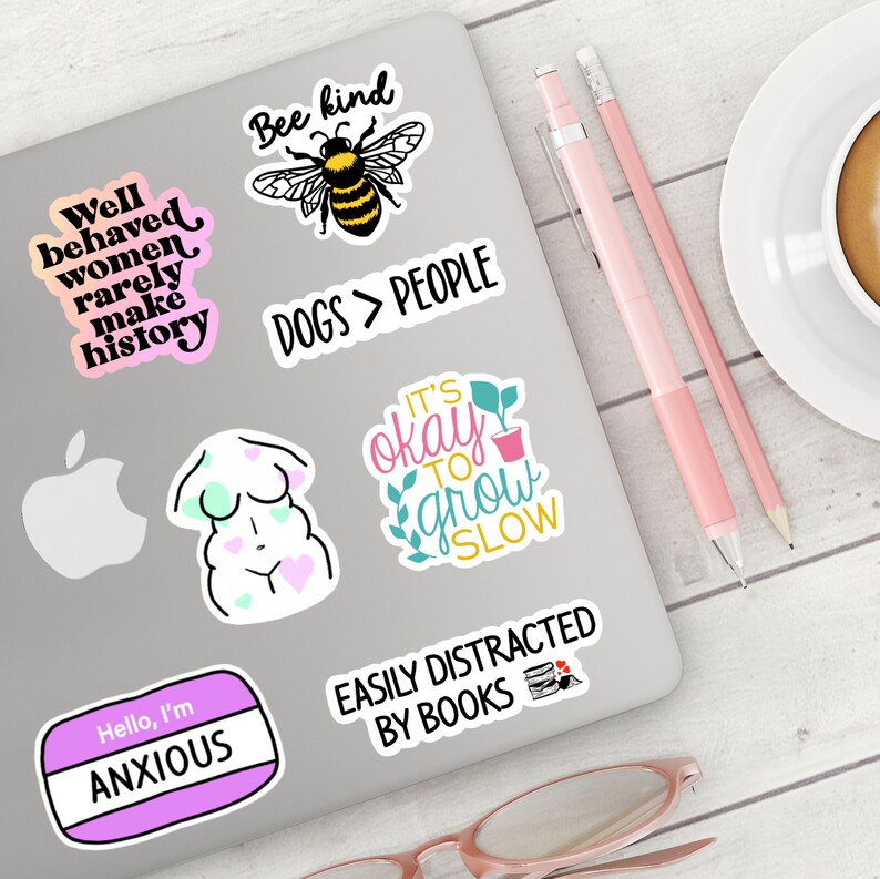 May include: A laptop computer with various stickers on it. The stickers feature phrases like "Well behaved women rarely make history", "Bee kind", "Dogs > People", "It's okay to grow slow", "Easily distracted by books", and "Hello, I'm Anxious".