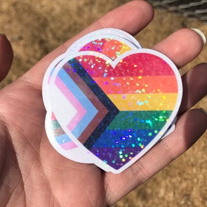 May include: Three heart-shaped stickers with a rainbow, transgender, and bisexual pride flag design. The stickers are iridescent and have a glitter finish.