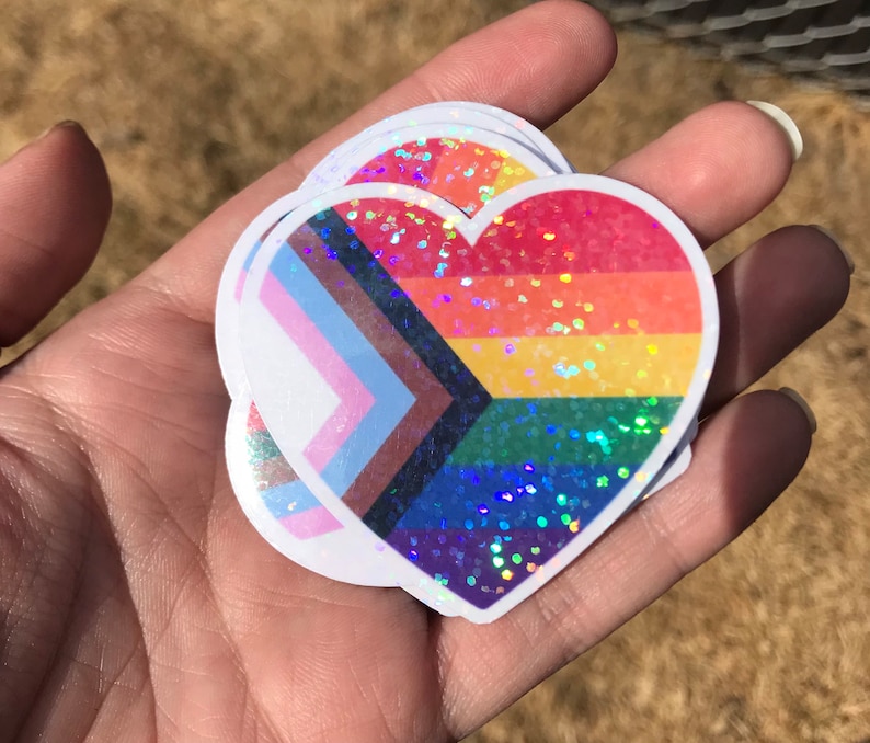 May include: Three heart-shaped stickers with a rainbow, transgender, and bisexual pride flag design. The stickers are iridescent and have a glitter finish.
