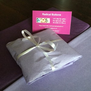 May include: A purple gift box wrapped in tissue paper and tied with a white ribbon. The box is sitting on a purple surface. A business card for Radical Buttons is visible in the background. The card says "Let's make the world a less oppressive place, one button at a time :)" and includes a website address and email address.
