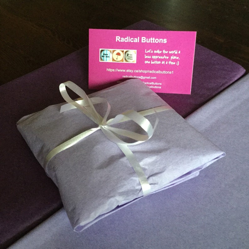 May include: A purple gift box wrapped in tissue paper and tied with a white ribbon. The box is sitting on a purple surface. A business card for Radical Buttons is visible in the background. The card says "Let's make the world a less oppressive place, one button at a time :)" and includes a website address and email address.