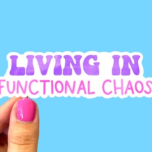 May include: A sticker with the text "LIVING IN FUNCTIONAL CHAOS" in purple and pink lettering. The sticker is held by a hand with pink nail polish against a light blue background.