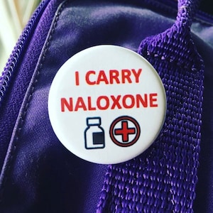Könnte beinhalten: Weißer Knopf mit dem Text "I CARRY NALOXONE" in roten Buchstaben. Unter dem Text befindet sich ein rotes Kreuz und eine schwarz-weiße Illustration einer Flasche.