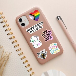 May include: A pink phone case with various stickers. The stickers include a rainbow heart, a body positivity message, a bee with the words "Bee kind", a plant with the words "It's okay to grow slow", and a text sticker that says "Well behaved women rarely make history".