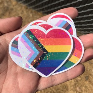 May include: A set of five heart-shaped stickers with a rainbow, transgender, and bisexual pride flag design. The stickers are iridescent and have a glitter finish.