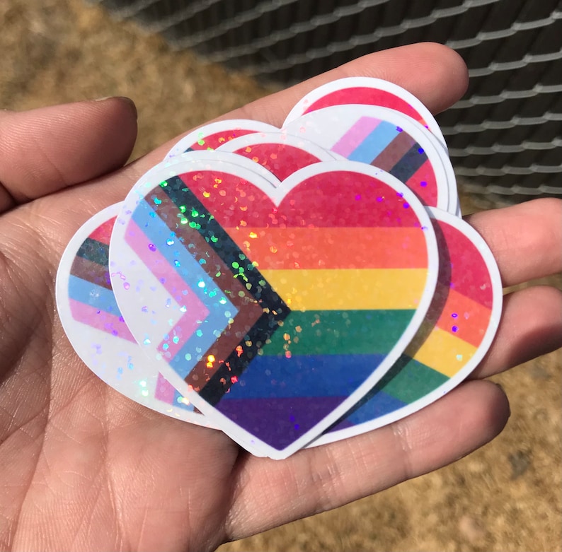 May include: A set of five heart-shaped stickers with a rainbow, transgender, and bisexual pride flag design. The stickers are iridescent and have a glitter finish.