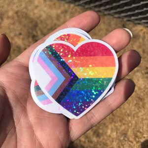 May include: A stack of heart-shaped stickers with a rainbow and transgender pride flag design. The stickers are covered in iridescent glitter.
