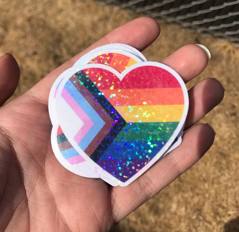 May include: A stack of heart-shaped stickers with a rainbow and transgender pride flag design. The stickers are covered in iridescent glitter.