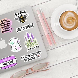 May include: A laptop covered in stickers, including a bee with "Bee kind" text, a lavender plant, and the phrase "Dogs > People." A cup of coffee, pencils, and glasses are also present.