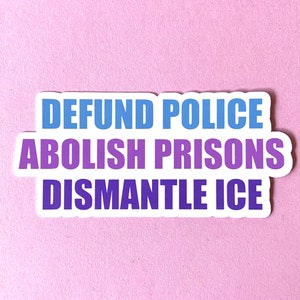 May include: A white sticker with the text "DEFUND POLICE, ABOLISH PRISONS, DISMANTLE ICE" in blue and purple lettering. The sticker is on a light pink background.