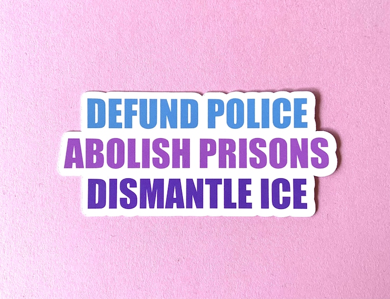 May include: A white sticker with the text "DEFUND POLICE, ABOLISH PRISONS, DISMANTLE ICE" in blue and purple lettering. The sticker is on a light pink background.