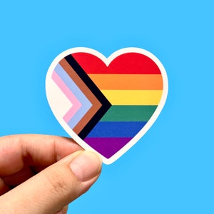 May include: A heart-shaped sticker with a rainbow flag design, including black, brown, light blue, pink, yellow, orange, red, green, blue, and purple stripes. The flag is a symbol of inclusivity and diversity.