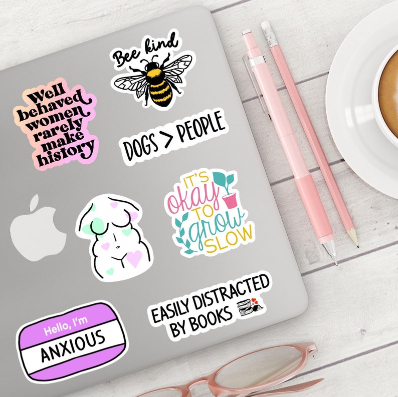Puede incluir: Un ordenador port&aacute;til con varias pegatinas coloridas. Las pegatinas presentan diferentes frases e im&aacute;genes, incluyendo "Well behaved women rarely make history", "Bee kind", "Dogs > People", "It's okay to grow slow", "Easily distracted by books", y "Hello, I'm Anxious".