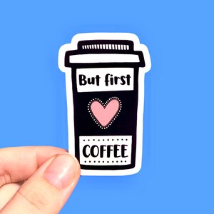 May include: A white sticker with a black outline of a coffee cup. The cup has the text "But first, coffee" and a pink heart.