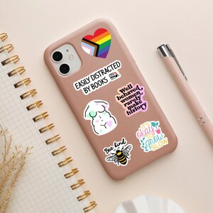 May include: A pink phone case with various colorful stickers. The stickers feature a rainbow heart, a woman's body, a bee, and text that reads "Easily Distracted by Books", "Well behaved women rarely make history", "Bee kind", and "It's okay to grow slow".