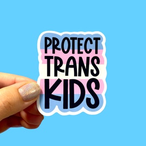 May include: A sticker with the text "PROTECT TRANS KIDS" in black, set against a background of the transgender pride flag colors: light blue, pink, and white. The sticker is held against a light blue background.