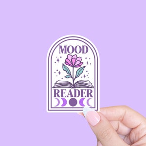 Könnte beinhalten: Ein weißer Aufkleber mit lila Rand und den Worten "MOOD READER" in einer stilisierten Schriftart. Der Aufkleber zeigt eine rosa Blume, die aus einem offenen Buch wächst, mit Mondphasen darunter. Der Hintergrund ist helllila.