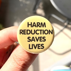 May include: A yellow button with the text "Harm Reduction Saves Lives" in black lettering.