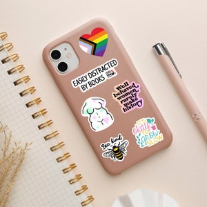 Puede incluir: Una funda de tel&eacute;fono rosa con varias pegatinas de colores. Las pegatinas muestran un coraz&oacute;n arco&iacute;ris, un cuerpo de mujer, una abeja y texto que dice "F&aacute;cilmente distra&iacute;do por los libros", "Las mujeres bien portadas rara vez hacen historia", "S&eacute; amable" y "Est&aacute; bien crecer despacio".