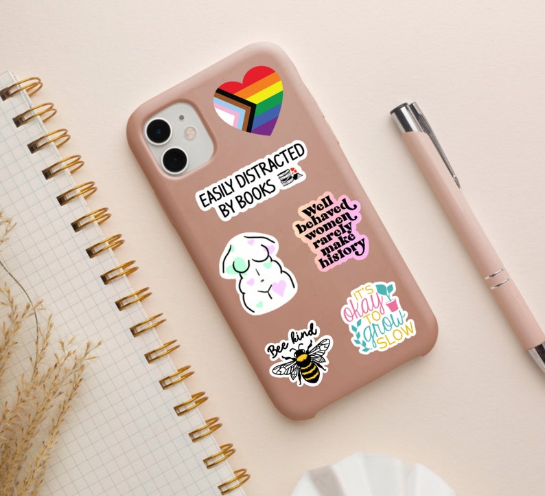 Puede incluir: Una funda de tel&eacute;fono rosa con varias pegatinas de colores. Las pegatinas muestran un coraz&oacute;n arco&iacute;ris, un cuerpo de mujer, una abeja y texto que dice "F&aacute;cilmente distra&iacute;do por los libros", "Las mujeres bien portadas rara vez hacen historia", "S&eacute; amable" y "Est&aacute; bien crecer despacio".