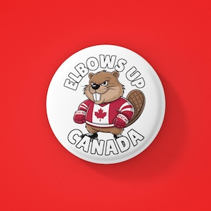 May include: White button featuring a cartoon beaver wearing a red and white hockey jersey with a Canadian flag on the front. The text "Elbows Up Canada" is written around the beaver.