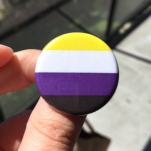 May include: A round button featuring the non-binary pride flag. The flag has four horizontal stripes: yellow, white, purple, and black.