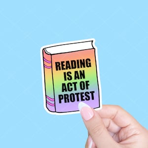 May include: A sticker in the shape of a book with a rainbow gradient spine and cover. The text "READING IS AN ACT OF PROTEST" is printed in black on the cover. The sticker is held by a hand against a light blue background.