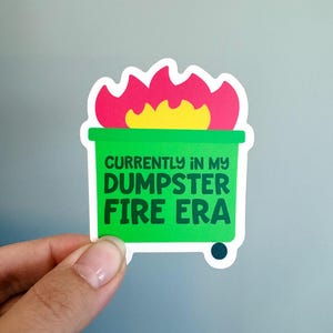 May include: A die-cut sticker featuring a bright green dumpster with a vibrant pink and yellow flame graphic emerging from the top. The text printed in dark green on the front of the dumpster reads, "CURRENTLY IN MY DUMPSTER FIRE ERA."