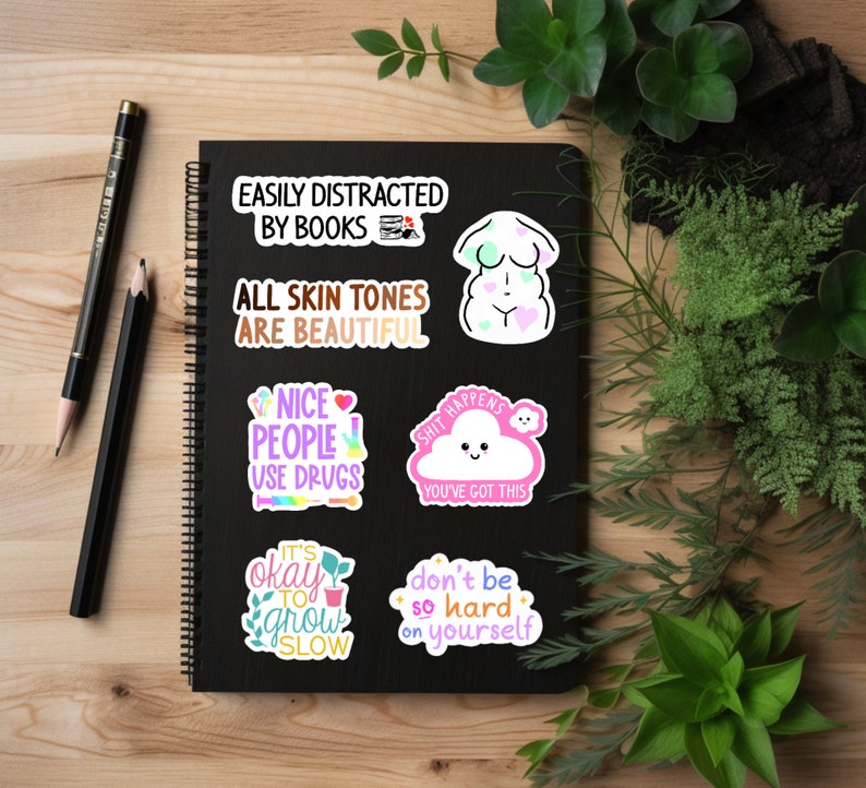 May include: A black spiral-bound notebook with several colorful stickers. The stickers have various phrases, including "EASILY DISTRACTED BY BOOKS," "ALL SKIN TONES ARE BEAUTIFUL," and "SHIT HAPPENS YOU'VE GOT THIS." Two pencils and greenery surround the notebook.