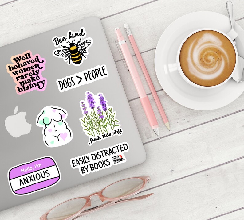 May include: A laptop covered in stickers, including a bee with "Bee kind" text, a lavender plant, and the phrase "Dogs > People." A cup of coffee, pencils, and glasses are also present.