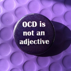 Puede incluir: Un bot&oacute;n redondo, morado oscuro, con el texto blanco "OCD is not an adjective". El bot&oacute;n est&aacute; sobre un fondo morado claro texturizado, con una sombra proyectada.