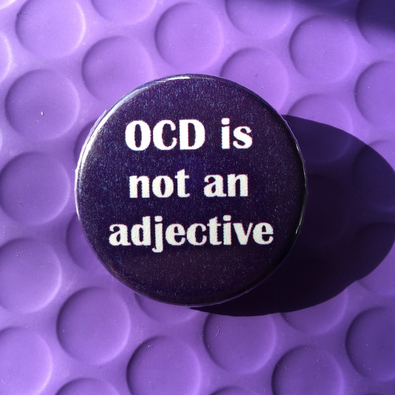 Puede incluir: Un bot&oacute;n redondo, morado oscuro, con el texto blanco "OCD is not an adjective". El bot&oacute;n est&aacute; sobre un fondo morado claro texturizado, con una sombra proyectada.