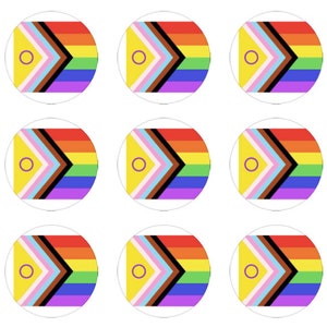 May include: Nine circular stickers with a rainbow flag design. The flag has a yellow circle with a purple ring in the center, followed by a pink stripe, a light brown stripe, a dark brown stripe, a black stripe, a yellow stripe, a green stripe, a light blue stripe, and a purple stripe.