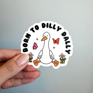 May include: A white die-cut sticker featuring a cartoon goose with an orange beak and feet. The text BORN TO DILLY DALLY is printed in black around the goose, accompanied by two small pink and orange butterflies, two pink flowers, and stars.