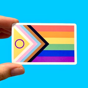 May include: A rectangular sticker featuring the Progress Pride flag design. The flag includes the traditional rainbow stripes, along with additional colors and symbols representing inclusivity. The sticker is held against a light blue background.