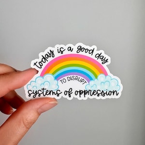 Peut inclure: Un autocollant découpé représentant un arc-en-ciel coloré au-dessus de nuages bleu clair. Le texte Today is a good day est écrit en noir au-dessus, TO DISRUPT en lettres capitales noires, et systems of oppression en écriture noire en dessous.
